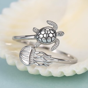 Peut inclure: Une bague ouverte argentée présentant un motif de tortue de mer à une extrémité et un motif de méduse à l'autre. La carapace de la tortue présente un motif en grille détaillé et la méduse possède de longs tentacules fluides.