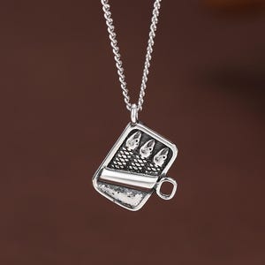 Puede incluir: Un collar con colgante plateado. El colgante es un dije rectangular detallado con un diseño de taza. El dije tiene un asa pequeña y patrones intrincados, incluyendo tres pequeñas formas redondas. La cadena del collar es un diseño delicado y enlazado.