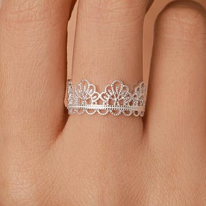 May include: A silver ring featuring an intricate, openwork lace-like pattern that resembles a delicate crown. The band has a scalloped edge and a detailed filigree design, crafted with a polished metallic finish that catches the light.