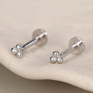 May include: Two silver-colored stud earrings with a cluster of three small spheres at the end of each post. The earrings are displayed on a light-colored, slightly textured surface. The earrings have a flat, round backing.