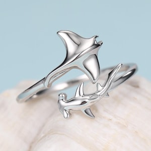 May include: A silver-toned open ring featuring a manta ray design on one end and a hammerhead shark design on the other. The metal band is polished and reflective, showcasing the distinct shapes of the two marine creatures meeting in the centre.