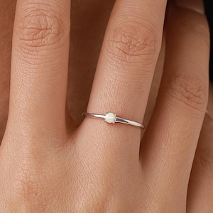 Könnte beinhalten: Ein zarter Silberring mit einem kleinen, runden, weißen Edelstein. Der Ring wird an einem Finger gezeigt, was sein minimalistisches Design und seine subtile Eleganz hervorhebt. Das Band ist dünn und poliert, was einen raffinierten Look erzeugt.