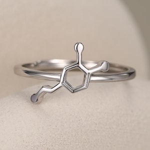 Puede incluir: Un anillo de tono plateado que presenta un motivo de estructura química central. El diseño consiste en un anillo hexagonal conectado a una corta cadena de átomos, todo en un acabado metálico pulido con una banda lisa y continua.