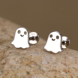 May include: A pair of silver ghost-shaped stud earrings. Each earring features a smiling ghost design with a rounded body and a small post for piercing. The earrings are displayed on a textured, light brown surface.