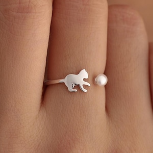 May include: A silver ring featuring a cat silhouette and a small pearl. The ring is worn on a finger, showcasing the delicate design. The cat is facing right, with its tail slightly curved.