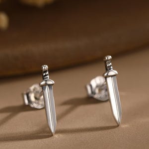 Op de afbeelding: Een paar zilverkleurige zwaardvormige oorstekers. De oorbellen zijn klein, met een gedetailleerd ontwerp van handvat en lemmet. De achtergrond is zachtbruin, met een wazig bloemelement in de linkerbovenhoek.