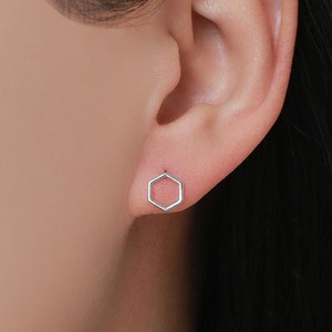 May include: A pair of small, silver-colored hexagon stud earrings. The earrings are simple in design, with a geometric shape. The earrings are shown on an ear, against a neutral background.