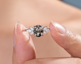 May include: A silver ring with a green and black oval gemstone surrounded by smaller clear gemstones. The ring is on a finger.