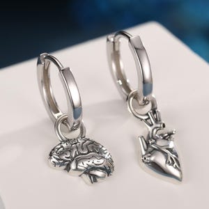 May include: Silver hoop earrings with dangling charms. One earring features a brain charm, the other a heart charm. The earrings are displayed on a white surface against a blue background. The earrings are made of a shiny metal.