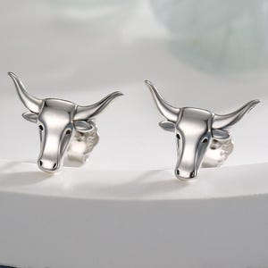 Puede incluir: Un par de pendientes de botón plateados con forma de cabeza de toro. Los pendientes se muestran sobre una superficie blanca sobre un fondo azul suave. Los pendientes tienen cuernos y rasgos faciales detallados.