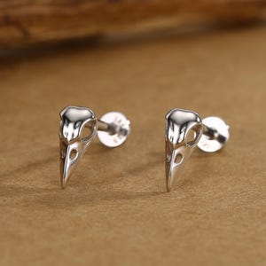 Puede incluir: Un par de pendientes de plata con forma de calaveras de pájaro. Los pendientes tienen un diseño detallado con cuencas de los ojos y picos visibles. El fondo es de un tono cálido y neutro, con un trozo de madera en la esquina superior izquierda.