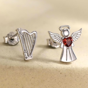 May include: A pair of silver stud earrings. One earring is a harp, the other is an angel with a red heart-shaped gemstone. The angel has wings and a halo. The earrings are on a neutral-toned surface.