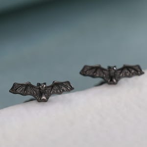 Puede incluir: Un par de pendientes de botón en forma de murciélago gris oscuro. Los pendientes tienen detalles de alas y cuerpo. Los pendientes se muestran sobre una superficie blanca con un fondo azul claro.
