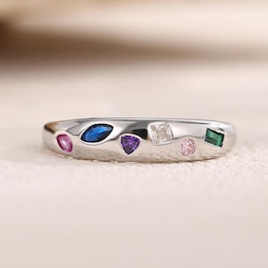 May include: A silver ring with a row of colorful gemstones. The ring features a blue oval, a purple triangle, a clear square, a pink round, and a green square gemstone.