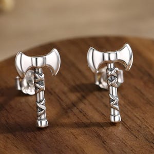 Puede incluir: Un par de pendientes de plata con forma de hacha. Los pendientes presentan un diseño detallado con una hoja curva y un mango texturizado. Los pendientes se exhiben sobre una superficie de madera.