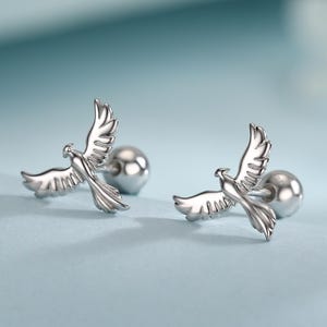 Puede incluir: Un par de pendientes de plata. Cada pendiente presenta un diseño detallado de paloma con las alas extendidas, posado sobre una pequeña base redonda. Los pendientes se exhiben sobre una superficie azul claro, con un fondo suave y borroso.