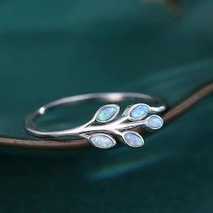 Puede incluir: Un anillo de plata con un delicado diseño de rama con cinco piedras de ópalo. El anillo está colocado sobre un fondo verde.