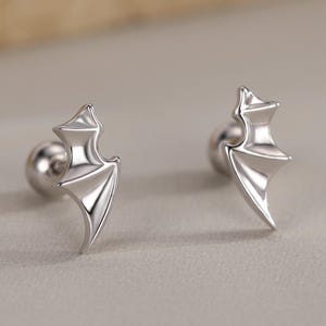 May include: A pair of silver stud earrings, each with a bat wing design. The earrings have a small, round backing. They are presented against a neutral backdrop, emphasizing their metallic shine.