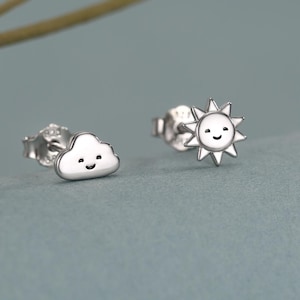 May include: A pair of silver stud earrings. One earring is a smiling cloud, and the other is a smiling sun. The earrings are on a blue-gray surface.