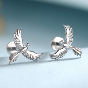 Puede incluir: Un par de pendientes plateados con forma de pájaro en vuelo. Los pendientes presentan alas detalladas y están sobre un fondo azul claro. Un diseño delicado y elegante, ideal para cualquier ocasión.