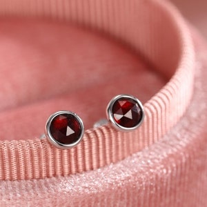 Pode incluir: Um par de brincos de prata com pequenas pedras redondas de cor vermelha.