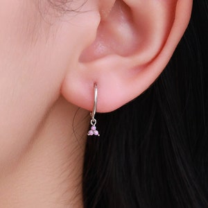 May include: A silver hoop earring with three small pink beads hanging beneath. The earring is worn on an ear, with the focus on the jewellery. The background is a soft blue.