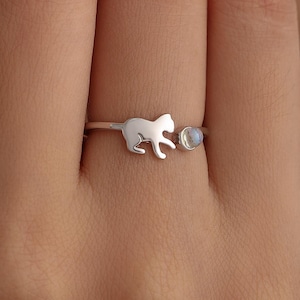 Op de afbeelding: Een zilveren ring met een katten-silhouet en een kleine, ronde, lichtgekleurde steen. De kat is in profiel afgebeeld, met een licht gebogen staart. De ring wordt aan een vinger gedragen, wat het delicate ontwerp benadrukt.