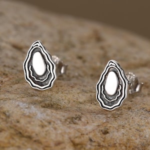 May include: A pair of silver stud earrings, each featuring an abstract, layered design with a white center. The earrings are set against a neutral, textured background. The earrings have a unique, organic shape.