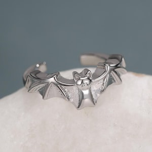 Puede incluir: Un anillo abierto de color plateado que presenta un motivo central de murciélago con las alas extendidas. La banda de metal es pulida y reflectante, mostrando la forma detallada del cuerpo y las alas del murciélago.