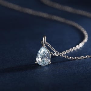 Può includere: Una collana color argento con un ciondolo a forma di pera in pietra preziosa azzurra, sospeso a una catenina sottile. Il ciondolo è fissato da una piccola montatura in metallo. Nell'angolo è presente un piccolo logo bianco con le lettere SRS.