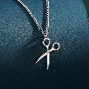 May include: A silver necklace featuring a scissors pendant. The pendant is a detailed miniature of scissors, suspended from a fine chain. The necklace is set against a dark teal backdrop.