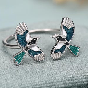 May include: An open-style silver ring featuring two bird motifs facing each other. Each bird is detailed with black, white, and teal enamel on the wings and tail feathers, creating a symmetrical design that wraps around the finger.