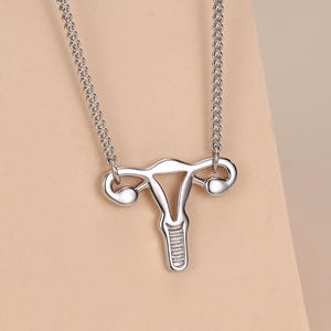 May include: A silver-colored necklace featuring a stylized anatomical uterus pendant. The pendant is suspended from a delicate chain, showcasing the shape of the uterus. The necklace is displayed against a neutral background.