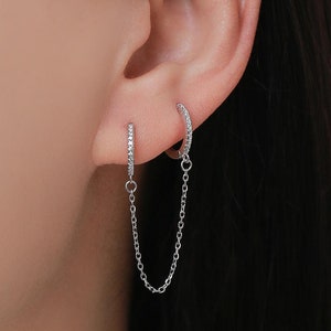 May include: A pair of silver earrings connected by a delicate chain. The earrings feature small, sparkling stones set along the hoops, creating a subtle yet elegant design. The earrings are shown on an ear, highlighting their delicate size and style.