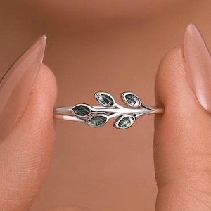 Puede incluir: Un anillo de plata con un diseño de hojas. El anillo tiene una banda delgada y está adornado con varias piedras preciosas ovaladas de color verde, que se asemejan a hojas. El anillo se sostiene entre dos dedos sobre un fondo borroso.