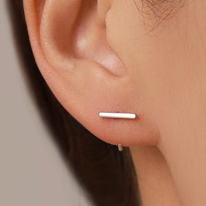 Puede incluir: Un pendiente de botón con forma de barra plateada que presenta un diseño rectangular horizontal. El pendiente está colocado en el lóbulo de la oreja, mostrando un acabado metálico liso y pulido que se asienta sobre la piel con una línea limpia.