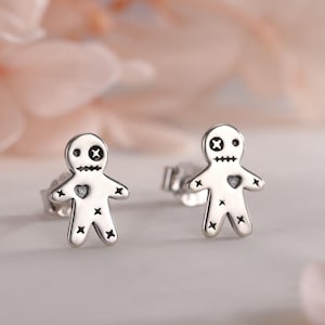 May include: A pair of silver-colored stud earrings shaped like voodoo dolls. Each doll has a heart-shaped chest, stitched limbs, and a stitched face with crossed eyes and a stitched mouth. The earrings are displayed on a white surface with blurred pink flowers in the background.