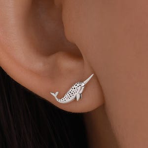 May include: A silver narwhal stud earring is shown on an ear. The earring is a small, detailed depiction of a narwhal, with a long tusk and dotted pattern on its body. The earring is worn on the ear.