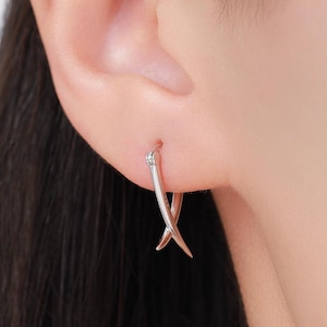 以下が含まれることがあります： X字型のデザインのシルバーのイヤリングのクローズアップ。イヤリングは耳に装着されています。イヤリングは光沢のある金属製です。イヤリングは小さく繊細なジュエリーです。