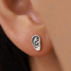Op de afbeelding: Een zilverkleurige oorbel in de vorm van een menselijk oor. De oorbel is klein en gedetailleerd, met de rondingen en contouren van een oor. De oorbel wordt gedragen op het oor van een persoon.