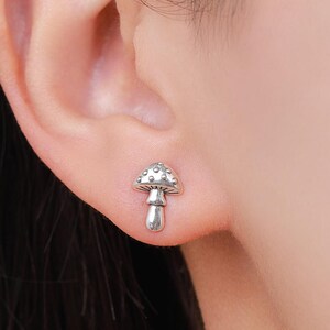 May include: A silver mushroom stud earring. The earring is a miniature mushroom design with a textured cap and a smooth stem. The earring is worn on an ear.