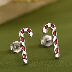 May include: A pair of silver candy cane stud earrings with red and white stripes. The earrings are on a green surface, with a blurred background. The earrings are a festive holiday accessory.