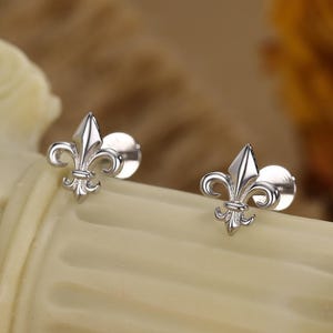 May include: A pair of silver-colored stud earrings, each featuring a fleur-de-lis design. The earrings are displayed on a cream-colored surface, with a blurred background of neutral tones and a hint of a dried flower. The earrings are small and delicate.