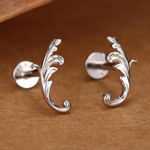 May include: A pair of silver stud earrings featuring a curved, ornate leaf-like design with intricate detailing. Each earring has a flat circular backing and a post, showcasing a polished metallic finish that reflects light across the curved surface.