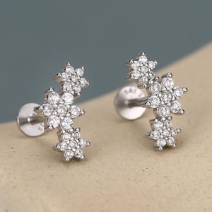 Peut inclure: Une paire de boucles d'oreilles à clous argentées. Chaque boucle d'oreille présente un groupe de petits cristaux clairs et étincelants disposés en forme de fleur. Les boucles d'oreilles sont posées sur un fond neutre.