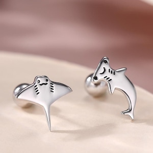 May include: A pair of silver stud earrings. One earring is shaped like a manta ray, the other like a dolphin. Both have a small, round post for piercing. The earrings are set against a soft, neutral background.