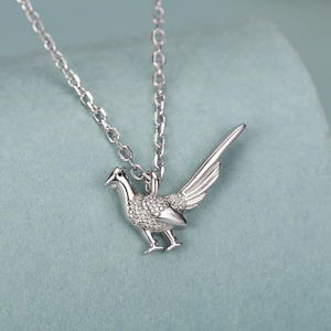 May include: A silver-colored necklace featuring a bird pendant with detailed wings and body. The bird is adorned with small, sparkling embellishments. The chain is delicate, and the pendant is centered against a soft, blue-green background.