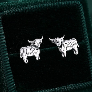 May include: A pair of silver earrings in the shape of a Highland cow, displayed in a green velvet box.