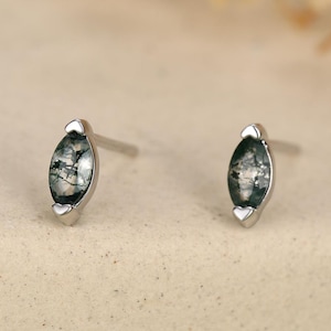 May include: A pair of silver stud earrings. Each earring features a marquise-shaped, moss agate stone set in a silver bezel. The earrings are displayed on a neutral surface with blurred floral accents in the background.
