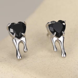 May include: A pair of stud earrings featuring a black heart-shaped gemstone set in a silver-toned metal base. The metal setting has a distinct dripping or melting design that extends downward from the base of the heart-shaped stone.